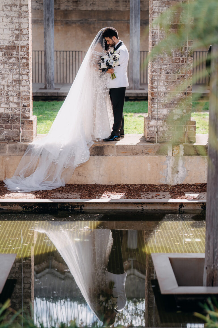 Bride and groom posing on a stone bridge with their wedding attire reflected in the water.