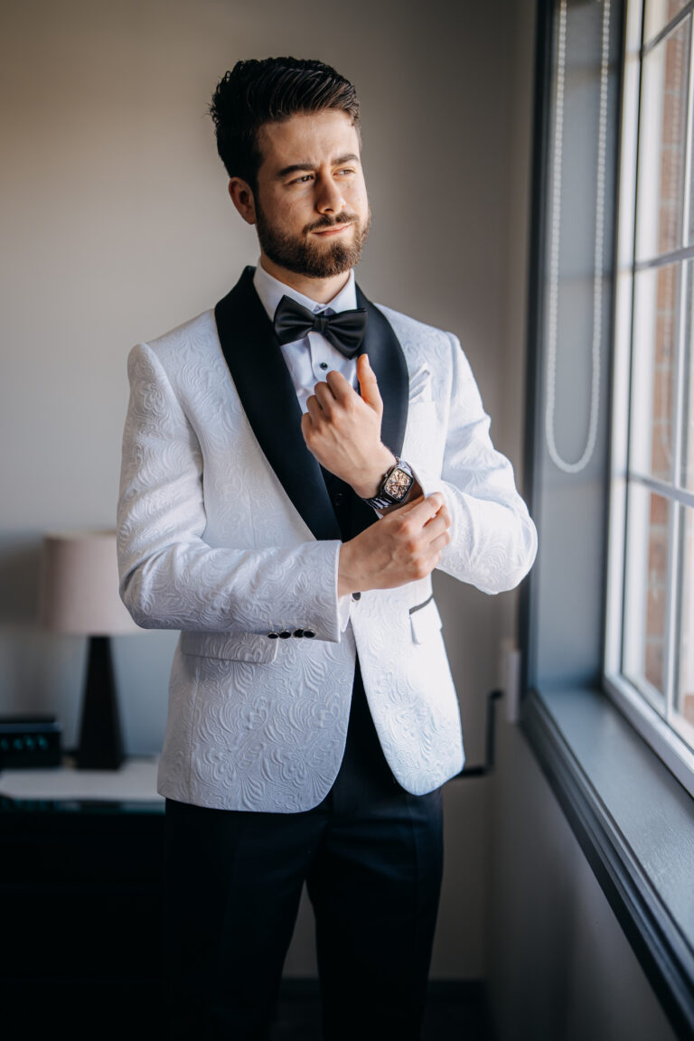 Groom Jordan adjusting his white tuxedo beside a window before his wedding ceremony.
