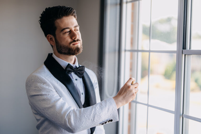 Natural light portrait of the groom wearing a white tuxedo beside a window.